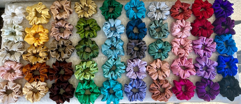 May include: A collection of 40 colourful satin scrunchies in various shades of green, blue, purple, brown, red, and white. The scrunchies are arranged in rows on a white surface.