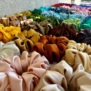 May include: A collection of colourful satin scrunchies in various shades of yellow, orange, brown, beige, and pink. The scrunchies are arranged in a row, with the colours gradually transitioning from yellow to pink.