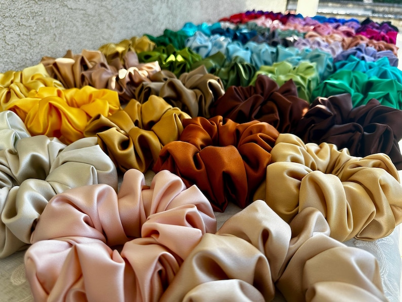 May include: A collection of colourful satin scrunchies in various shades of yellow, orange, brown, beige, and pink. The scrunchies are arranged in a row, with the colours gradually transitioning from yellow to pink.