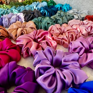 May include: A collection of colourful satin scrunchies in various shades of pink, purple, red, brown, green, and blue. The scrunchies are arranged in a semi-circle on a white surface.
