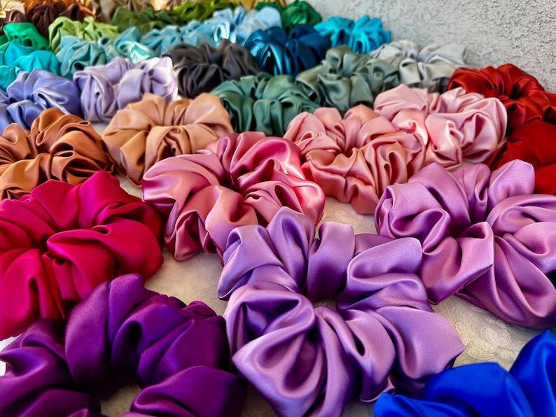May include: A collection of colourful satin scrunchies in various shades of pink, purple, red, brown, green, and blue. The scrunchies are arranged in a semi-circle on a white surface.