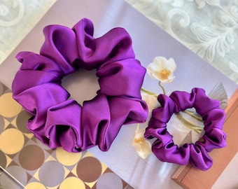 May include: Two purple satin scrunchies, one large and one small, on a patterned surface.