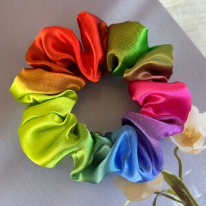 May include: A rainbow-coloured scrunchie made of satin fabric. The scrunchie is in a circle shape and has a gradient of colours, starting with red and ending with blue.