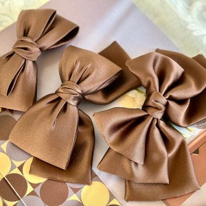 May include: Three brown satin hair bows of different sizes. The bows are arranged on a patterned surface.