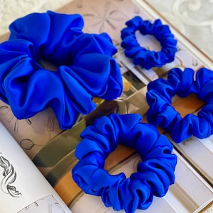 May include: Four blue satin scrunchies of different sizes. The scrunchies are arranged on a book with a white background.
