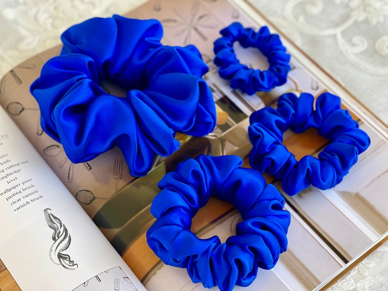 May include: Four blue satin scrunchies of different sizes. The scrunchies are arranged on a book with a white background.