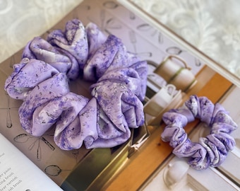 May include: Two purple and white patterned scrunchies. The larger scrunchie is in focus and the smaller scrunchie is out of focus in the background.