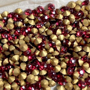 May include: A close-up shot of a pile of red and gold rhinestones. The rhinestones are a mix of red, faceted gems and gold, cone-shaped studs. The gems are scattered across a white surface, creating a sparkling effect.