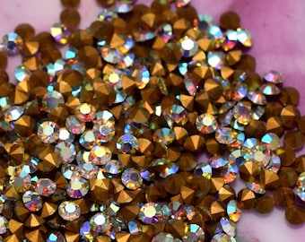 May include: A pile of small, round, bronze-colored rhinestones with iridescent facets. The rhinestones are scattered on a pink and white marbled surface, creating a sparkling effect. Ideal for crafts and embellishments.