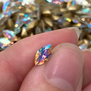 May include: A single iridescent marquise-shaped rhinestone with a pointed end. The rhinestone has a rainbow effect with shades of pink, blue, and green.