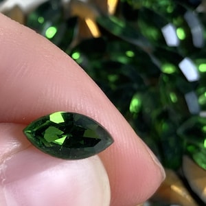 May include: A faceted, dark green gemstone in a marquise shape is held between fingers. The gemstone has a brilliant cut, reflecting light. Other similar gemstones are scattered in the background, some with a golden hue.