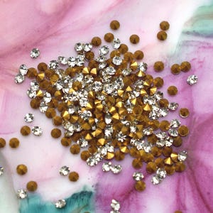 May include: A collection of gold and clear crystal rhinestones scattered on a pink and purple surface. The rhinestones are small, with a faceted design that catches the light. The gold rhinestones have a metallic finish.