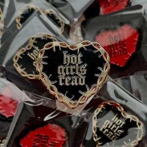 May include: A black and gold heart-shaped pin with barbed wire around the edge. The pin says "hot girls read" in gold lettering.