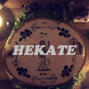 May include: A wooden circle with a black and white illustration of a woman holding a torch and surrounded by keys and snakes. The text "HEKATE" is written in bold white letters across the center of the circle.