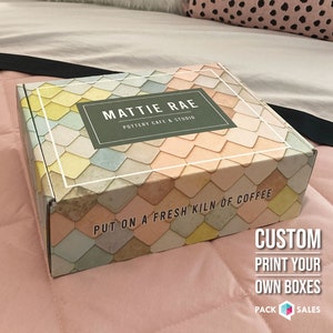 May include: A custom printed box with a colorful geometric pattern. The box has the text "MATTIE RAE POTTERY CAFE & STUDIO" and "PUT ON A FRESH KILN OF COFFEE". The text "CUSTOM PRINT YOUR OWN BOXES" is printed on a white background with a small box icon.