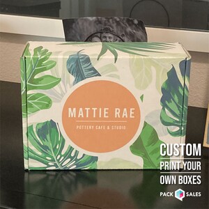 May include: A white box with a tropical leaf pattern and a peach-colored circle with the text "MATTIE RAE POTTERY CAFE & STUDIO". The box has the text "CUSTOM PRINT YOUR OWN BOXES PACK SALES" on the side.