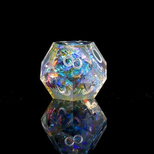 Puede incluir: Un dado D20 iridiscente multifacético con un exterior de resina transparente. El interior presenta un diseño colorido y fracturado con números y símbolos plateados. El dado se refleja en una superficie negra.