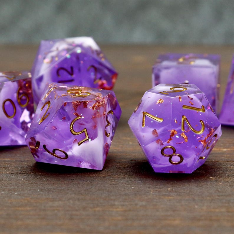 May include: Set of purple and white dice with gold flecks and numbers. The dice are made of resin and have a cloudy, translucent appearance.