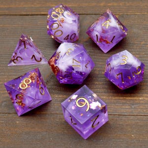 May include: Set of seven purple and white dice with gold flecks and numbers. The dice are made of resin and have a cloudy, swirled appearance.