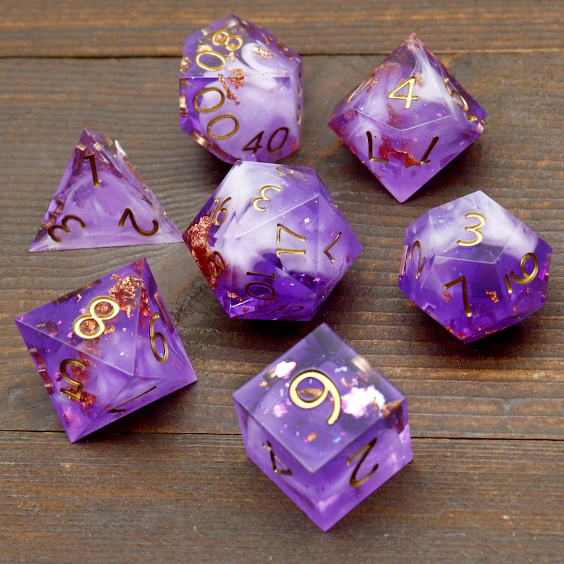 May include: Set of seven purple and white dice with gold flecks and numbers. The dice are made of resin and have a cloudy, swirled appearance.