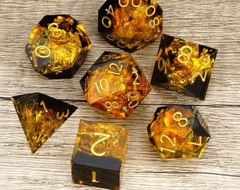 May include: A set of seven polyhedral dice featuring a translucent amber and black resin design with embedded gold foil flakes and clear gold-colored numerals. The collection includes a d20, d12, d10, d8, d6, d4, and a percentile die for gaming.