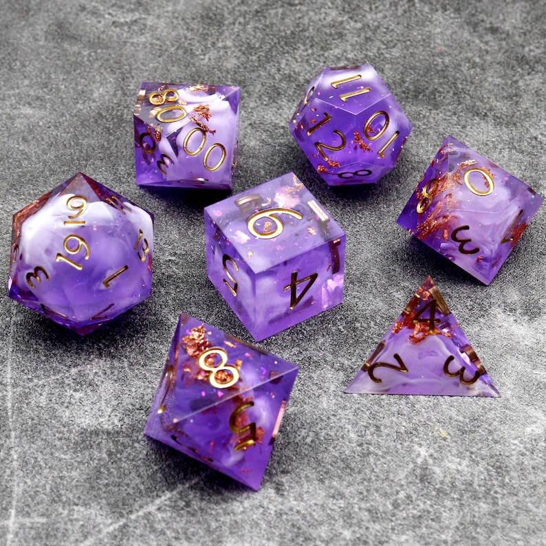 May include: Set of seven purple and white dice with gold flecks. The dice are a d4, d6, d8, d10, d%, d12, and d20.