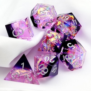 Op de afbeelding: Een set van zeven iriserende dobbelstenen met een paars en roze kleurenschema. De dobbelstenen zijn gemaakt van helder plastic met een holografische afwerking. De nummers op de dobbelstenen zijn zilver en zijn zichtbaar door het plastic. De dobbelstenen zijn verspreid over een wit oppervlak.