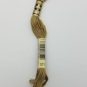 May include: A skein of olive green embroidery floss with a paper label featuring the DMC logo, the number 25, and the length 8 meters or 8.7 yards. The label also displays the color code 371 and a barcode, with text indicating sale outside the EU.