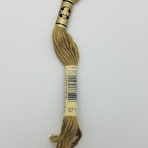 May include: A skein of DMC brand embroidery floss in a golden brown color. The label indicates it is size 25, 8 meters (8.7 yards) in length, and color number 371. The floss is tightly wound and ready for use in various needlework projects.