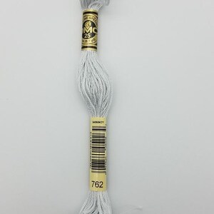 May include: A skein of light gray embroidery floss featuring a gold paper label with the text DMC, 25, and 8.7 yd. The label also displays a barcode, the number 762, and the code 5406042Y. The thread is wound into a long, twisted bundle.