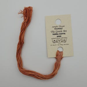May include: A length of terra cotta colored embroidery thread attached to a rectangular paper card. The card features the text Sampler Threads, The Gentle Art, TERRA COTTA, 0590, counted thread embroidery, and New Albany, Ohio 43054.