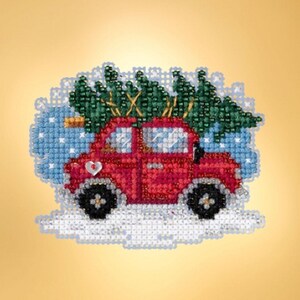 May include: A cross-stitch embroidery piece depicting a bright red car carrying a dark green Christmas tree on its roof. The car features black wheels, a small white heart detail on the side, and is set against a light blue background with white snowflakes.