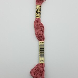 May include: A skein of dusty rose embroidery floss with a paper label featuring the DMC logo, the number 25, and the length of 8 meters (8.7 yards). The label also displays the color code 223 and a barcode for identification and inventory.