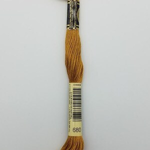 May include: A skein of mustard yellow embroidery floss made of 100% cotton. The thread is secured with two paper labels, one featuring the number 680, a barcode, and text indicating it is for sale outside the E.U. The other label lists material content.