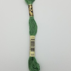 May include: A skein of sage green embroidery floss with a paper label featuring the DMC logo, the text 8m, a barcode, and the color number 320. The thread is wound into a long, twisted bundle, showcasing a consistent matte texture and color.
