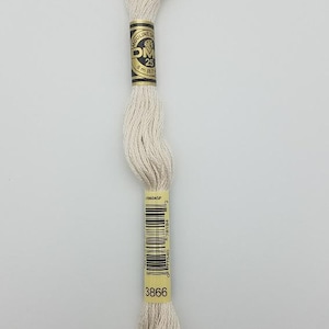 May include: A skein of off-white embroidery floss featuring a black and gold paper label with the text DMC, Mouliné, 25, 8 m (8.7 yd), and the number 3866 printed at the bottom, along with a barcode and the product code 1106045P.