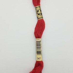 May include: A skein of deep red embroidery floss featuring a black and gold paper label with the text DMC, 25, and 8 m (8.7 yd). A second cream-colored label displays a barcode and the number 347, indicating the specific color shade.