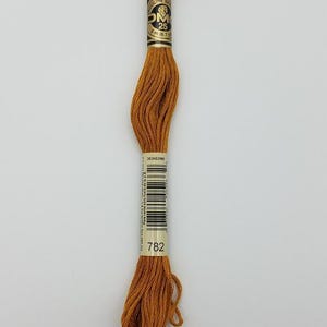 May include: A skein of golden-brown embroidery floss featuring a paper label with the text DMC, 25, 8 m (8.7 yd), and the color number 782. The thread is wound in a long, twisted bundle, displaying a consistent, rich, warm ochre color throughout.