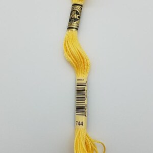 May include: A skein of bright yellow embroidery floss featuring a black and gold paper label with the text DMC, 25, 8.7 yd, and the number 744 printed on the bottom. The thread is wound in a long, twisted loop with loose ends at the bottom.