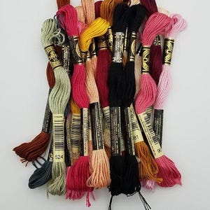 May include: A collection of DMC embroidery floss skeins in various colors including black, pink, yellow, green, and brown. Each skein features a paper label with the DMC logo, barcode, and specific color number, such as 524, wrapped around the thread.