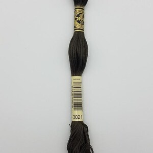 May include: A skein of dark brown embroidery floss with a gold and black paper label. The label features the text DMC, Mouliné Special, 25, 8 m (8.7 yd), and the color number 3021. The thread is wound into a long, twisted bundle.