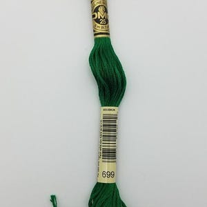 May include: A skein of vibrant green embroidery floss featuring a paper label with the text DMC, Mouliné Spécial, 25, 8 m (8.7 yd), and the color number 699. The thread is neatly wound and secured with a central band containing a barcode.