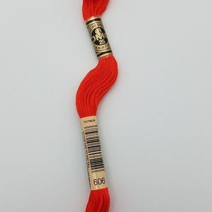 May include: A skein of bright red embroidery floss featuring a black and gold paper label with the text DMC, 25, 8 m (8.7 yd), 7527042R, and 606. The thread is wound in a long, twisted shape, showcasing a vibrant, solid color throughout the strand.