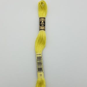 May include: A skein of bright yellow embroidery floss featuring a paper label with the text DMC, Moulin Special, 25, 8m (8.7 yd), 6018060F, and the number 11. The thread is wound in a long, slender loop, showcasing a vibrant, solid color.