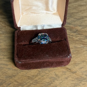 May include: A silver-toned ring featuring a square-cut, iridescent, multi-colored stone set in a prong setting. The band has a decorative lattice or cutout pattern along the sides. The ring is displayed inside a brown velvet-lined jewelry box.