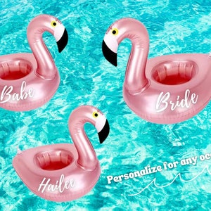 May include: Three pink inflatable flamingo drink holders with white text. The flamingo on the left says "Babe", the flamingo in the middle says "Hailee", and the flamingo on the right says "Bride". The flamingo drink holders are floating in a blue pool.