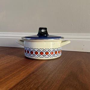 May include: A cream-coloured enamel pot with two side handles and a dark blue lid featuring a black knob. The exterior of the pot is decorated with a repeating geometric pattern in orange, white, and dark blue along the lower section of the body.
