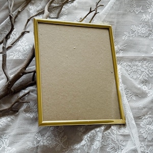 May include: A rectangular picture frame with a thin, metallic gold-coloured border and a plain, light brown cardboard backing. The frame is empty, showcasing a simple, clean design with straight edges and sharp corners, presented in a vertical orientation.