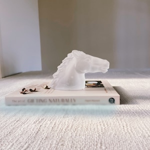 May include: A white glass horse head sculpture on top of a book titled "The art of Gifting Naturally" by Sagola Messard.