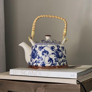 May include: A blue and white ceramic teapot with a floral design and a bamboo handle. The teapot is sitting on a white book with the word "SCANDI" printed on the cover.
